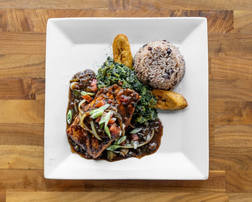 Grilled salmon with dark sauce, rice and beans, sauteed greens, and fried plantains on a white plate.