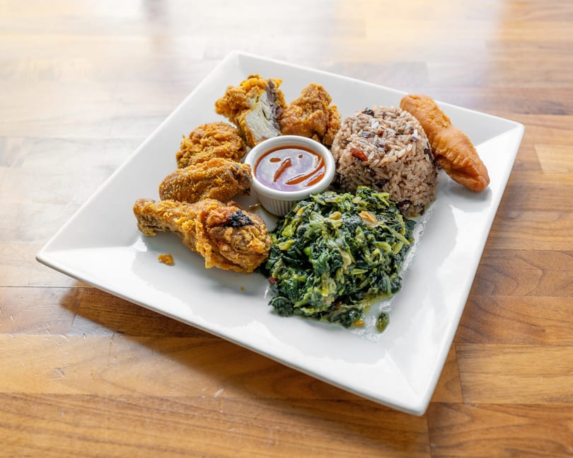 A white plate holds fried chicken, rice and peas, steamed callaloo, and a fried dumpling.