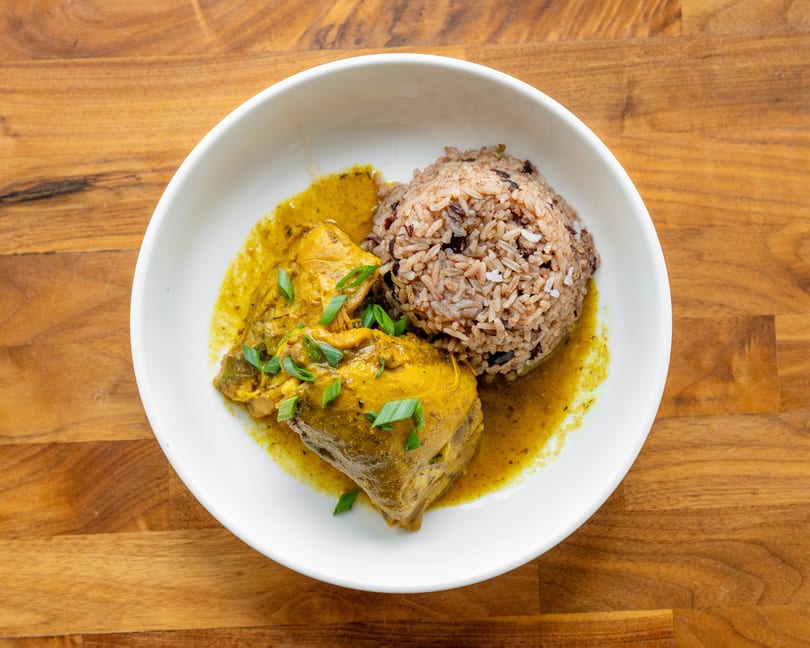 Curried chicken with rice and beans, garnished with green onions, in a white bowl on a wooden table.