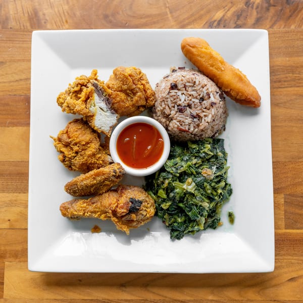Fried chicken pieces, rice and beans, collard greens, a fried plantain, and dipping sauce on a white plate.