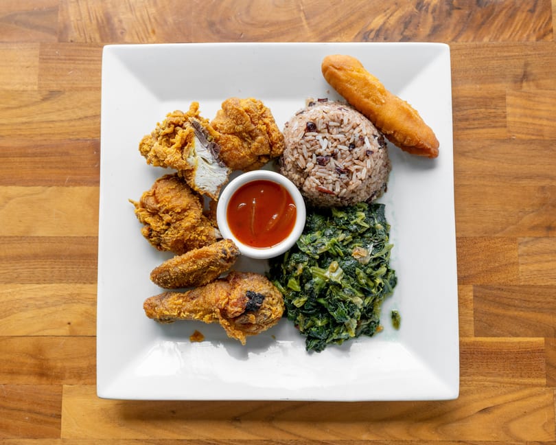 Fried chicken pieces, rice and beans, collard greens, a fried plantain, and dipping sauce on a white plate.
