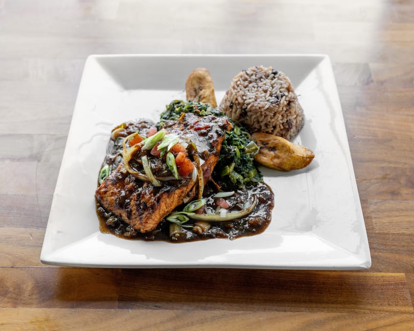 Grilled salmon with dark sauce, rice, greens, and plantains on a white plate.