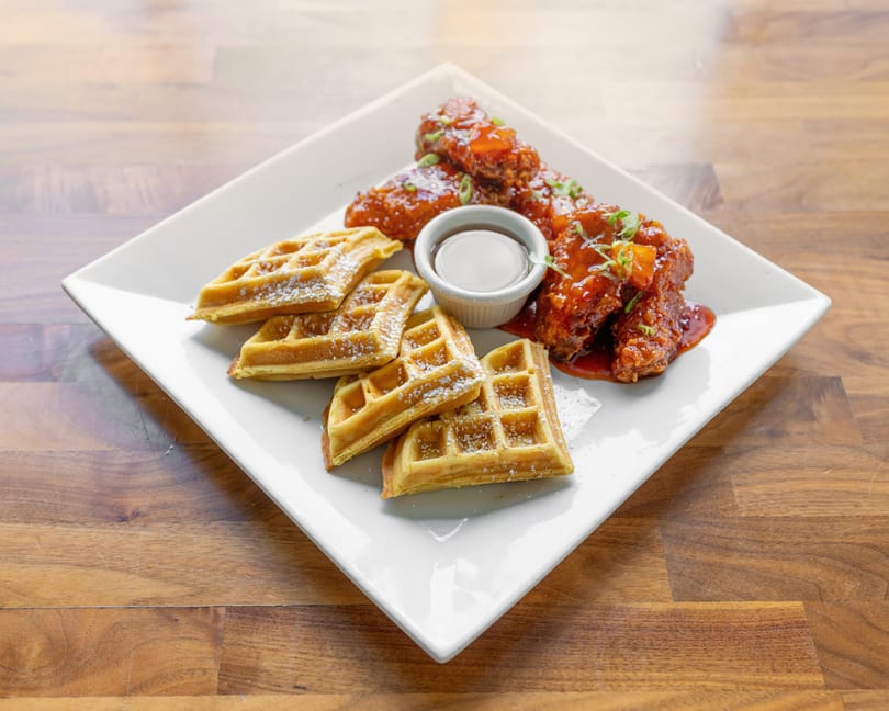 Chicken and waffles on a white plate, with syrup and powdered sugar, on a wooden table.