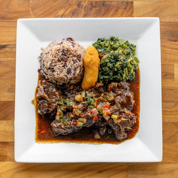 Oxtail stew with rice and peas, callaloo, and a fried dumpling on a white plate.