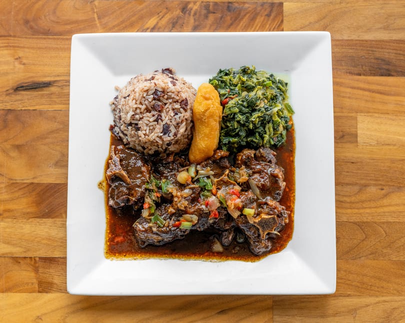 Oxtail stew with rice and peas, callaloo, and a fried dumpling on a white plate.