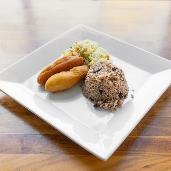 A white square plate holds two fried plantains, rice and beans, and coleslaw on a wooden table.