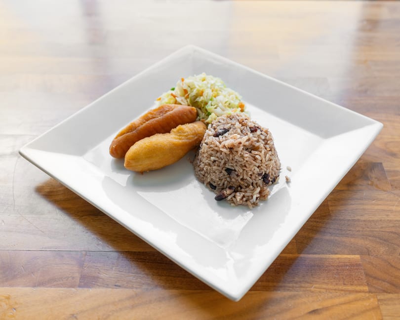 A white square plate holds two fried plantains, rice and beans, and coleslaw on a wooden table.