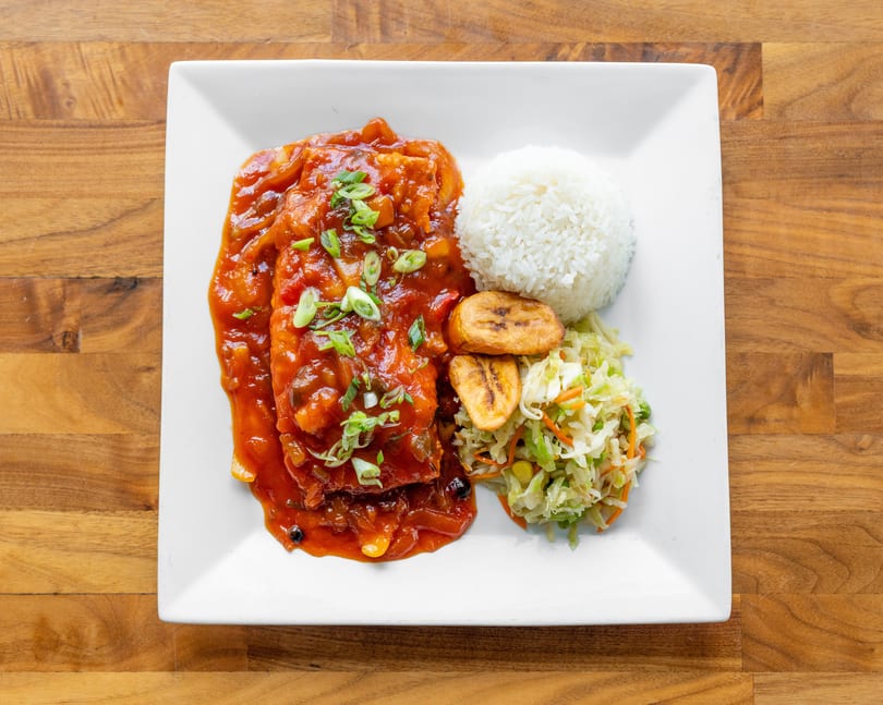 A white plate with fish in red sauce, white rice, fried plantains, and coleslaw.