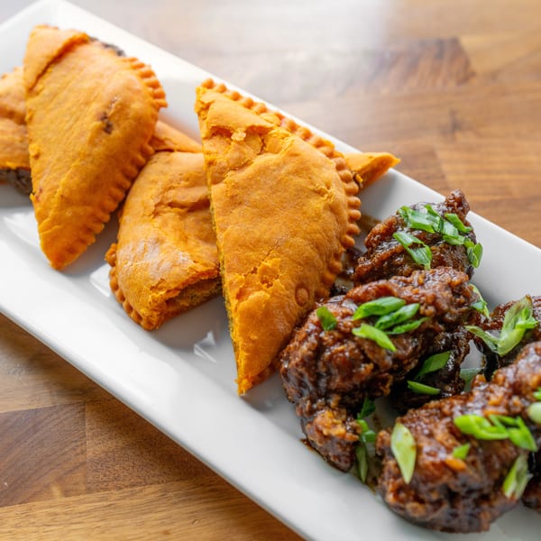 A white rectangular plate holds several golden-brown Jamaican patties and dark glazed chicken wings with scallions.