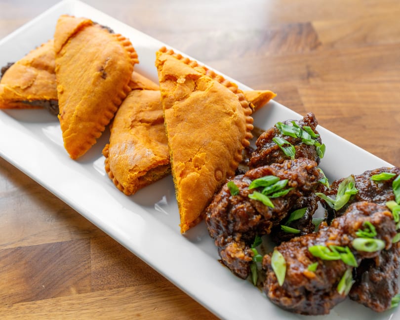 A white rectangular plate holds several golden-brown Jamaican patties and dark glazed chicken wings with scallions.