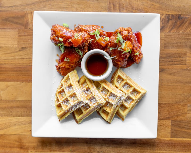 Chicken and waffles with syrup, garnished with green onions and diced fruit on a white plate.