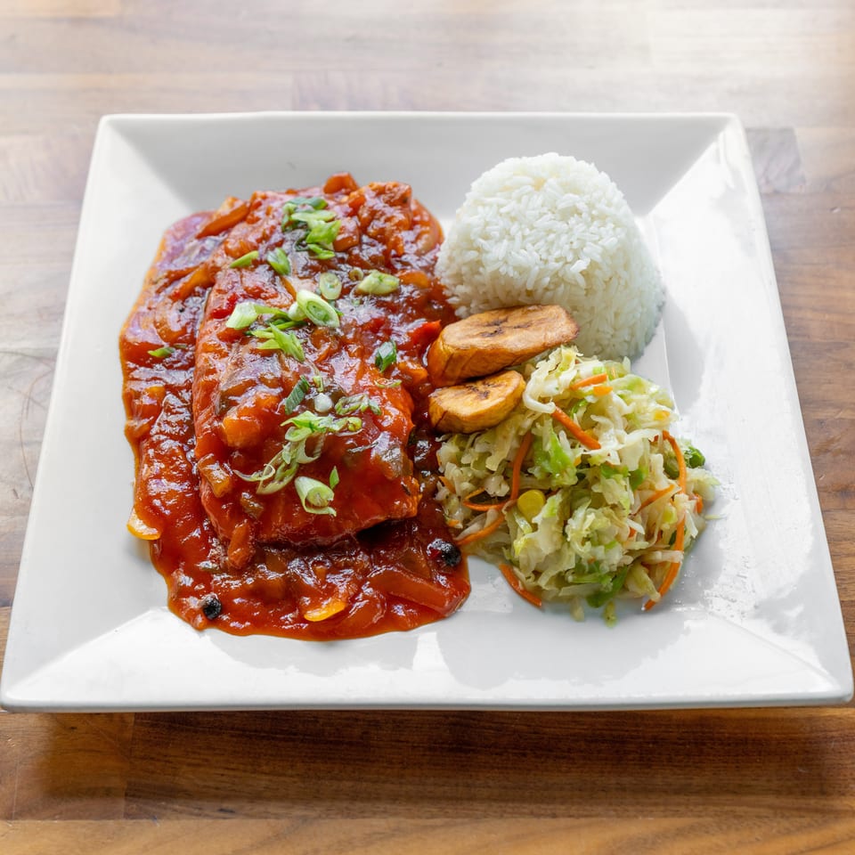 Two pieces of fish in red sauce with rice, fried plantains, and coleslaw on a white plate.
