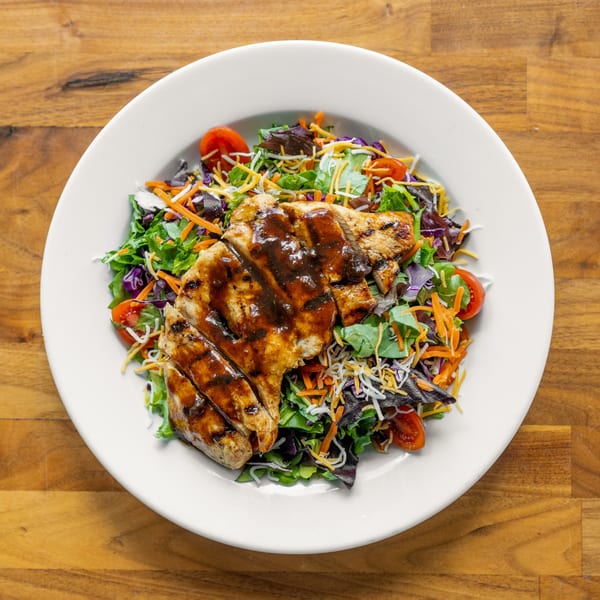 Grilled chicken salad with mixed greens, shredded carrots, cheese, and cherry tomatoes on a white plate.