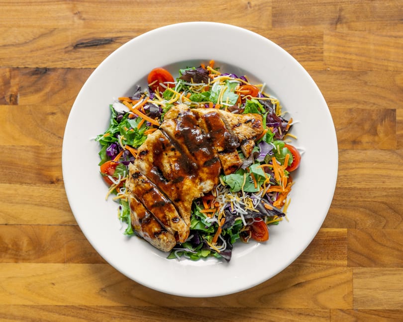 Grilled chicken salad with mixed greens, shredded carrots, cheese, and cherry tomatoes on a white plate.