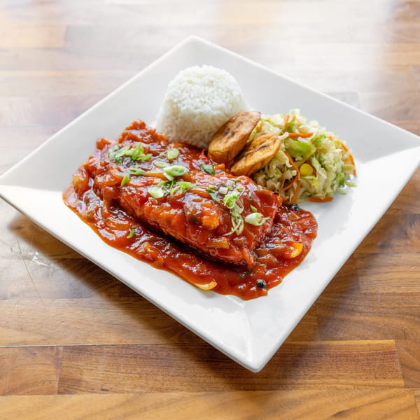 A white plate holds a fish fillet in red sauce, rice, fried plantains, and coleslaw.