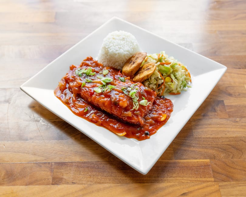 A white plate holds a fish fillet in red sauce, rice, fried plantains, and coleslaw.