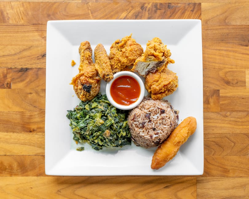 Fried chicken pieces, rice and beans, stewed greens, and a fried dumpling on a white plate.